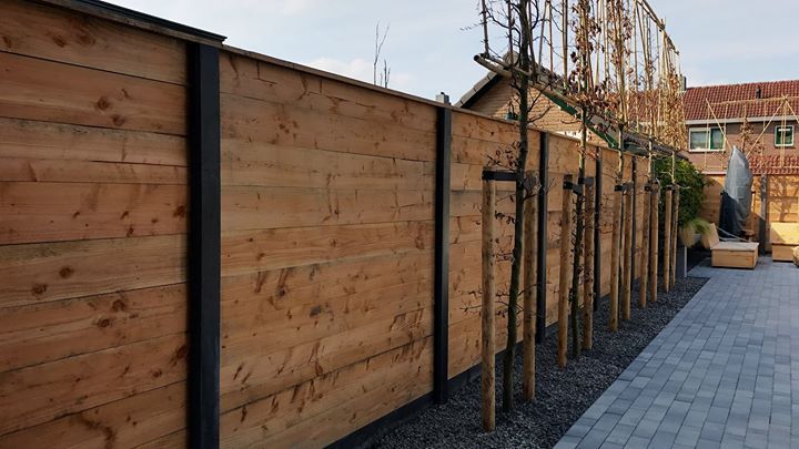 Houten schutting met donkere metalen palen, een rij jonge bomen langs een pad.