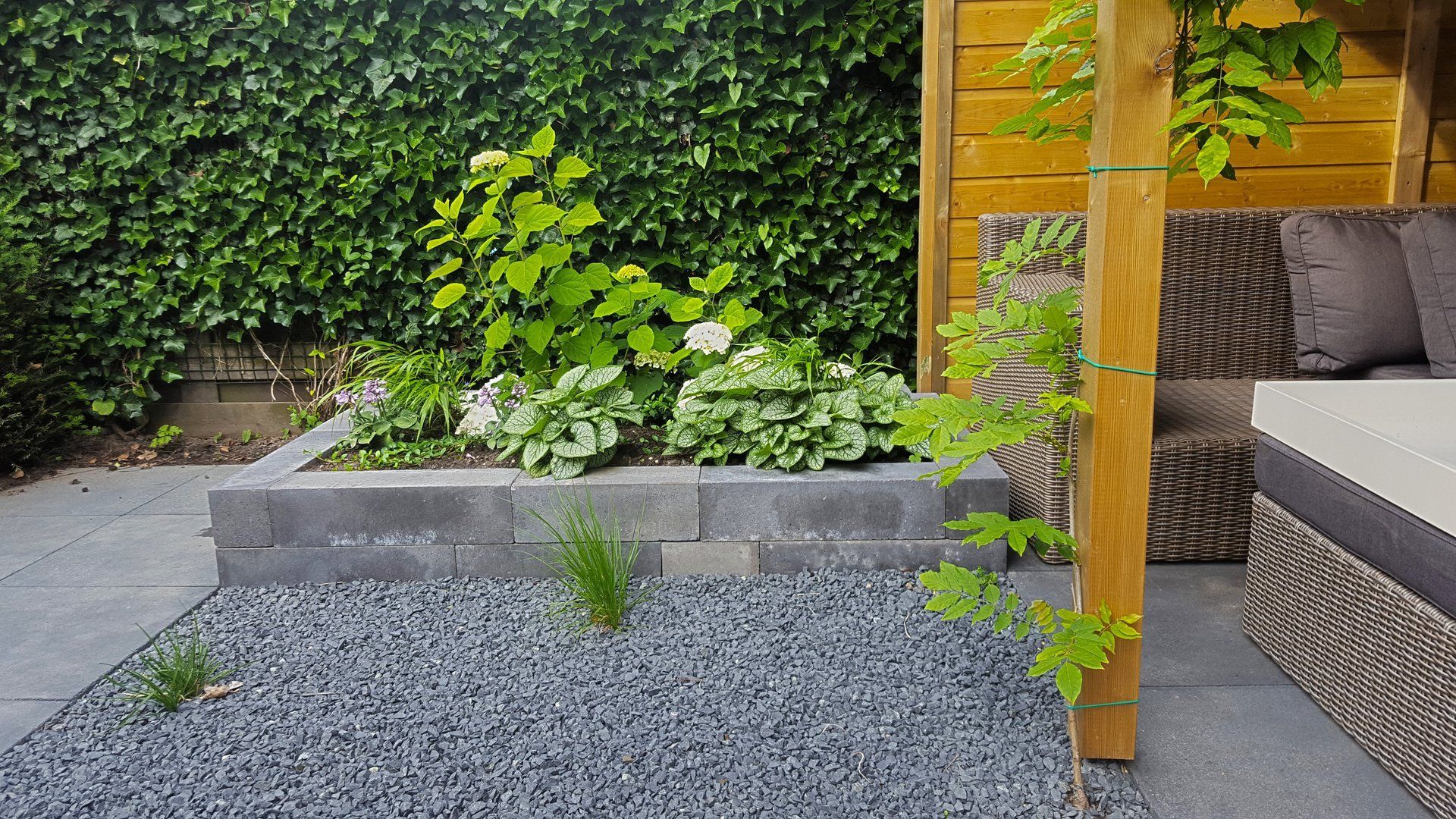 Een stenen tuinbed met weelderig groen, een grindpad en rieten tuinmeubelen onder een houten pergola.