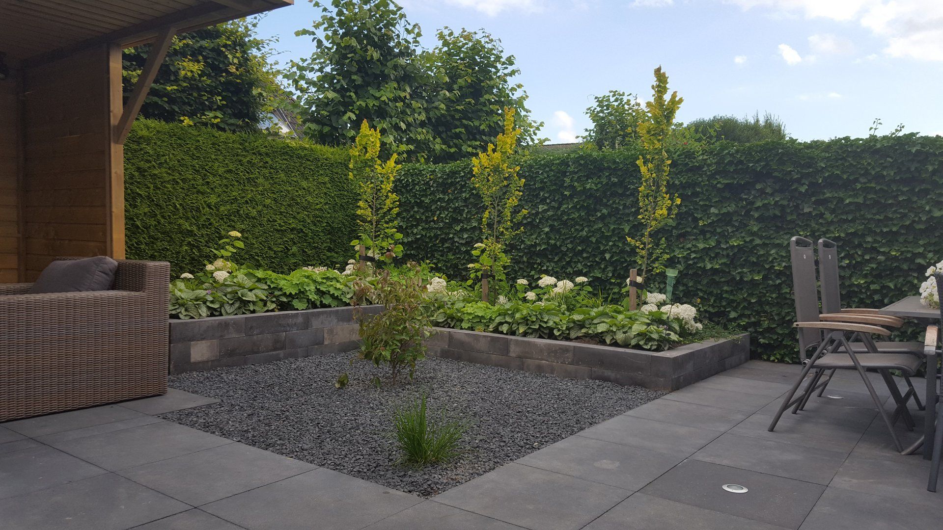 Terras met grijze tegels, verhoogde bloembedden en weelderige groene hagen.