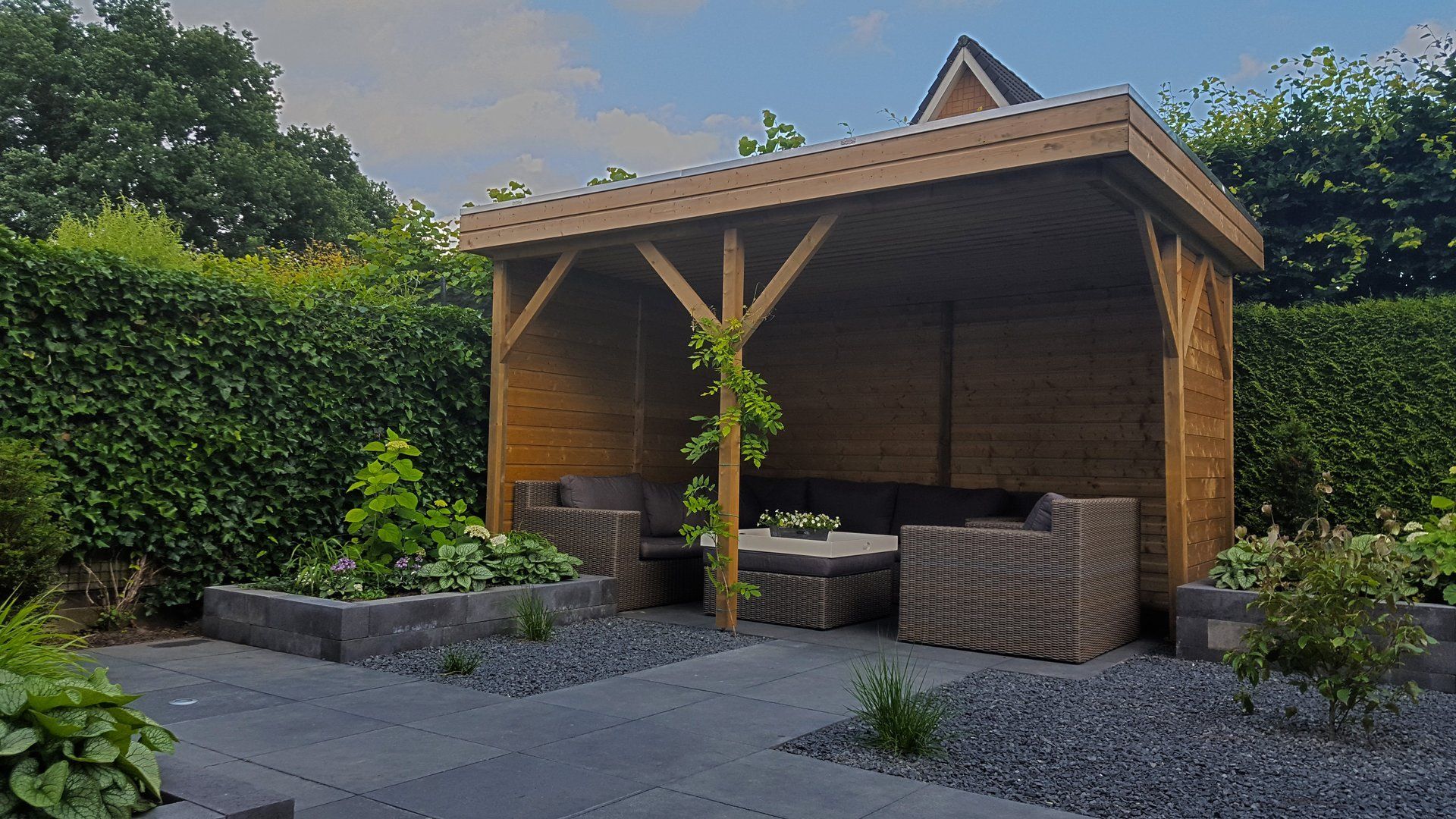 Houten prieel met zitgelegenheid in een tuin, omgeven door groen en donkergrijze stenen.