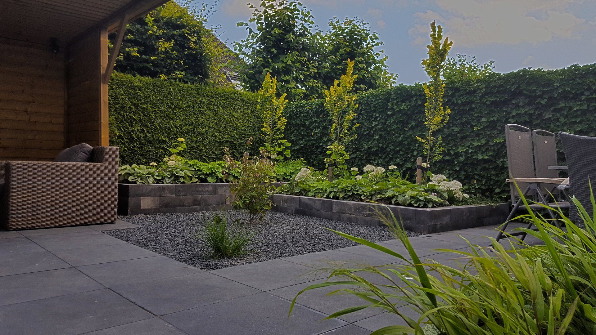 Terras met donkergrijze bestrating, verhoogde bloembedden en groen; een houten constructie aan de linkerkant.