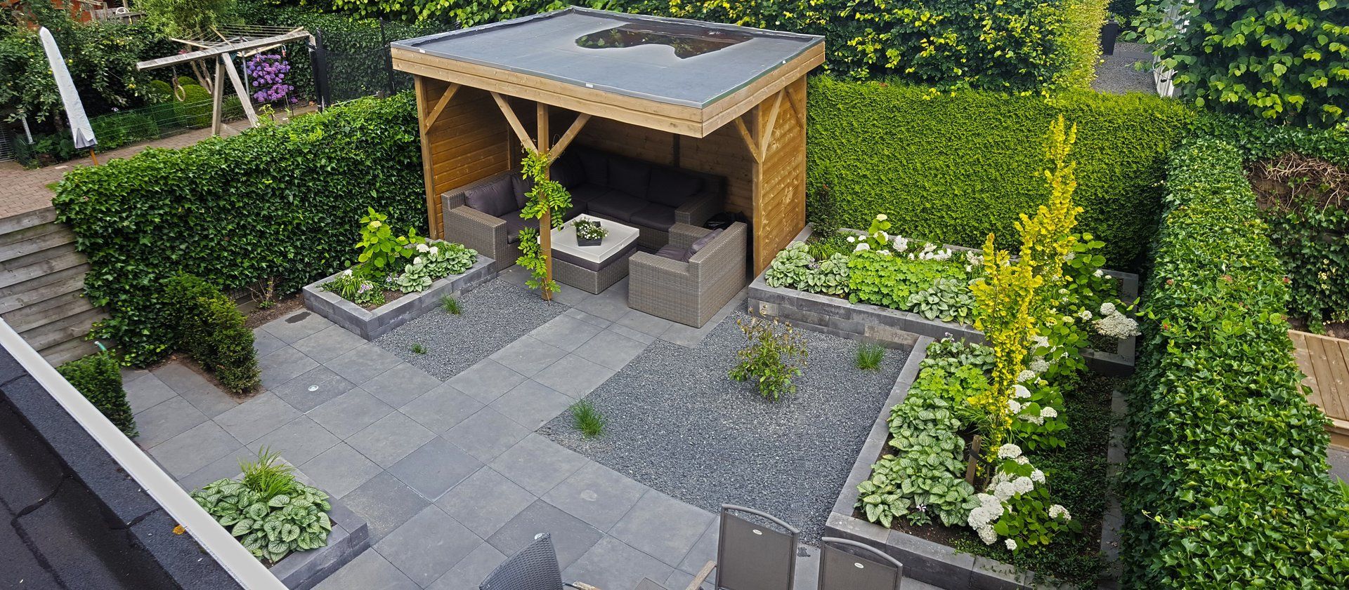 Bovenaanzicht van een moderne achtertuin met een houten prieel, stenen terras en groene hagen.