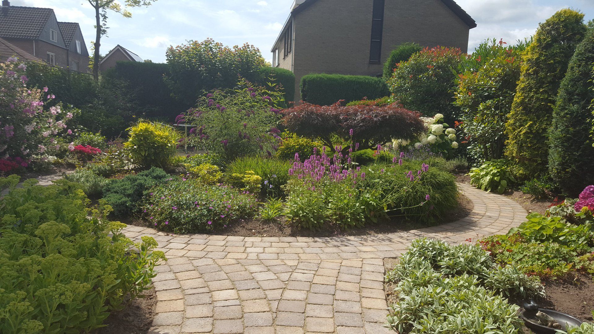 Een stenen pad door een weelderige tuin met diverse planten leidt naar een huis onder een bewolkte hemel.