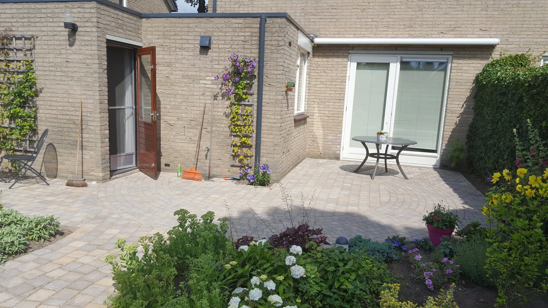 Terras in de achtertuin met bakstenen bestrating, een tafel en een schuifdeur, omgeven door bloembedden.