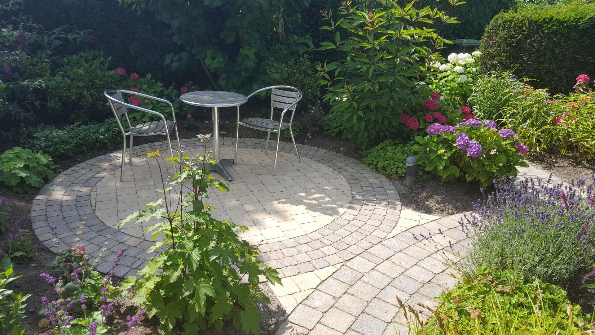 Rond stenen terras met tafel en stoelen, omgeven door een weelderige tuin.