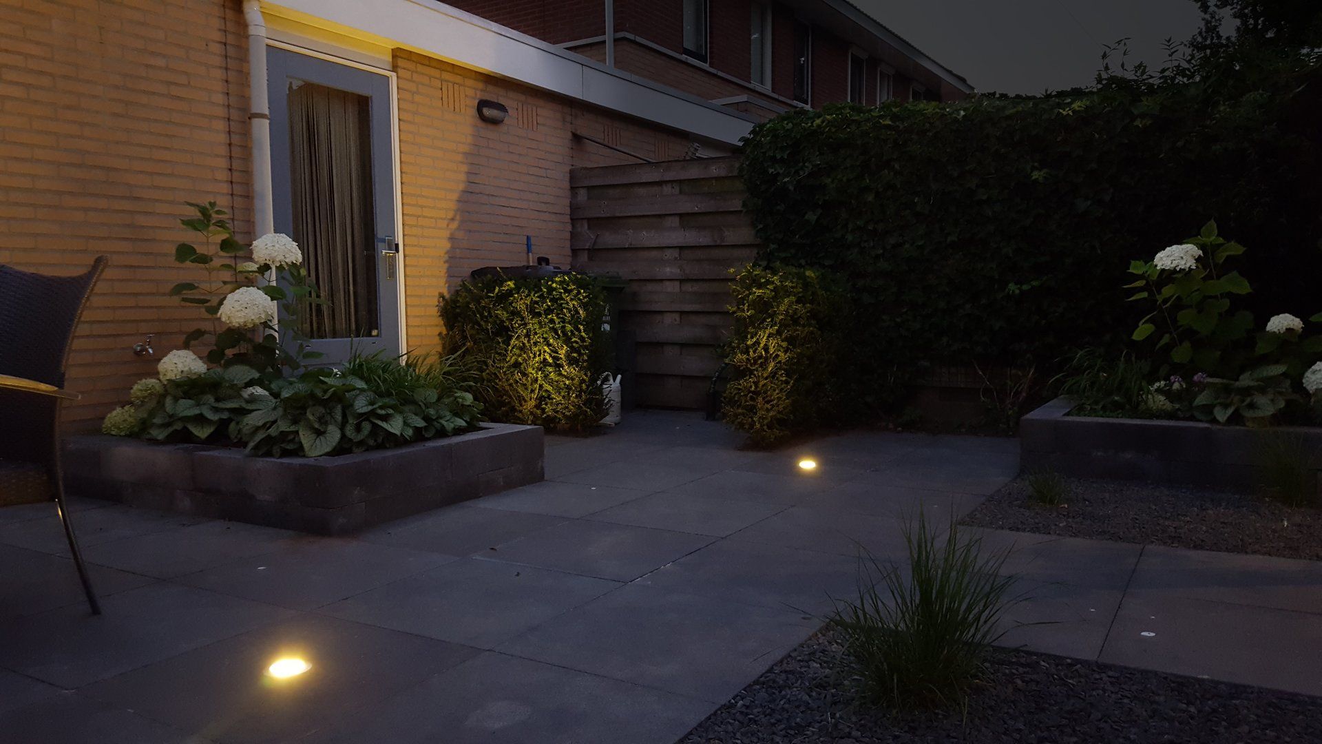 Terras in de achtertuin bij schemering, met verlichte bloemperken en grondverlichting. Bakstenen muur, houten schutting en weelderig groen.