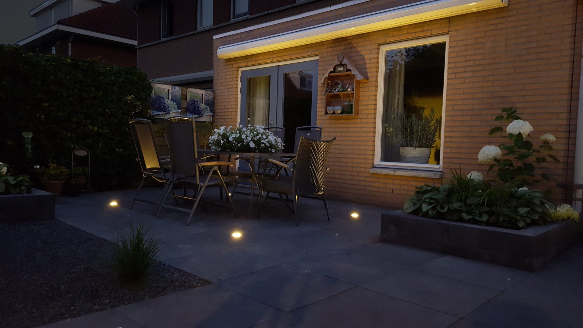 Terras bij schemering met tuinmeubilair, potplanten en grondverlichting die het geheel verlicht.