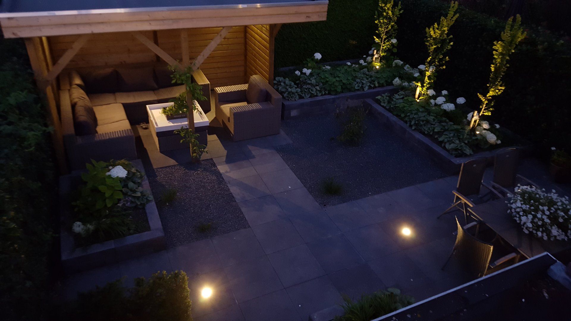 Een sfeervol terras in de achtertuin 's avonds met een verlicht prieel, bloemperken en buitenverlichting.