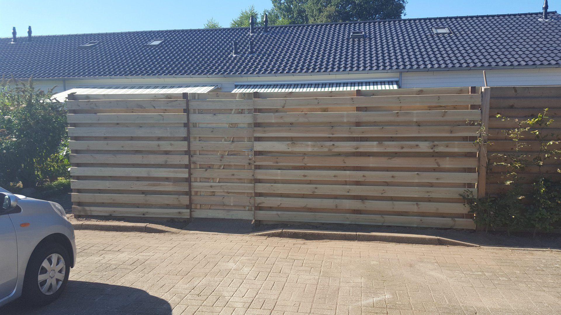 Houten schutting met horizontale latten. Grijs dak achter de schutting, geparkeerde zilverkleurige auto links.