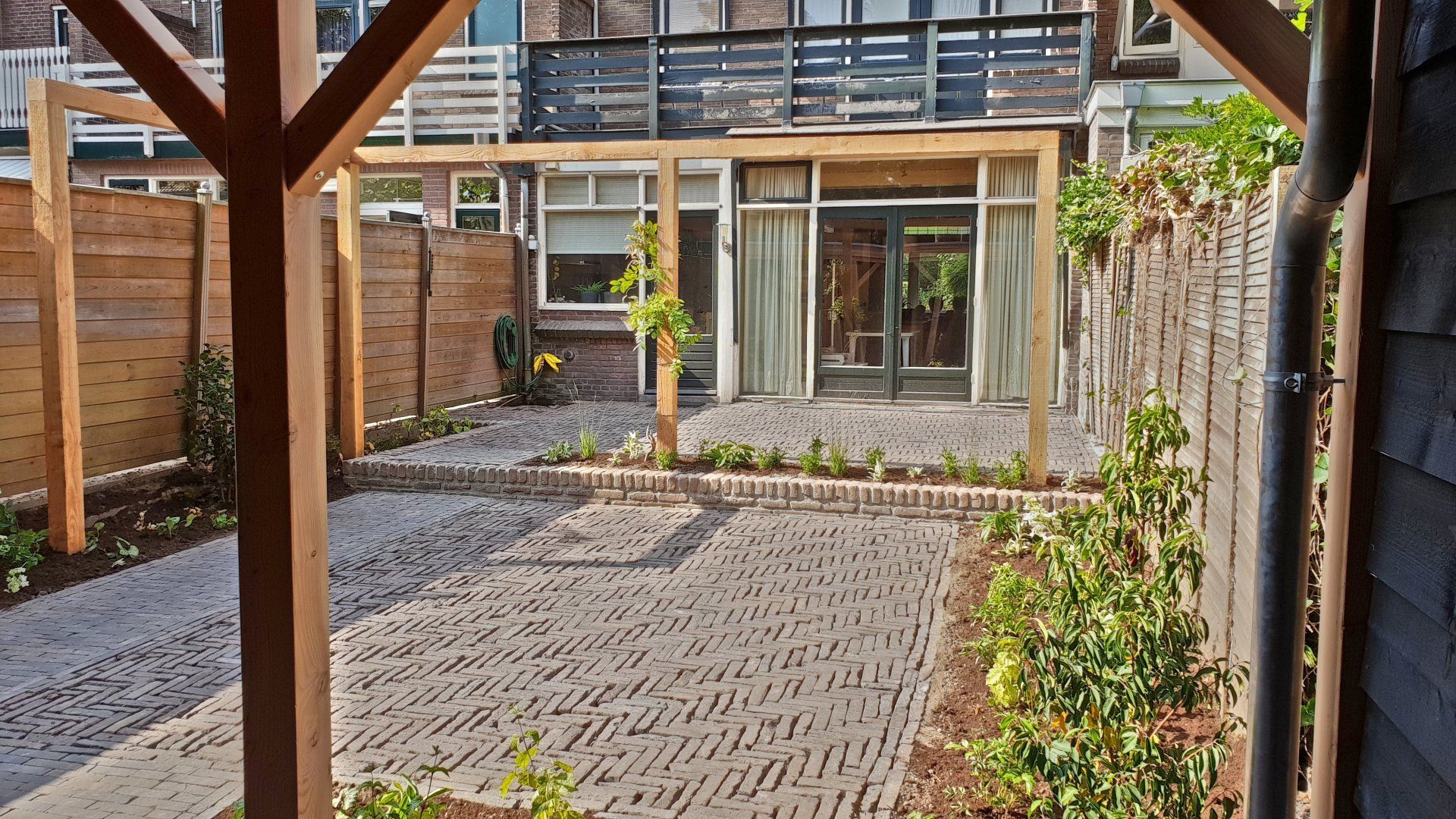 Achtertuin met stenen terras, houten pergola en schutting. Groene planten omzomen het terras. Huis met glazen deuren op de achtergrond.