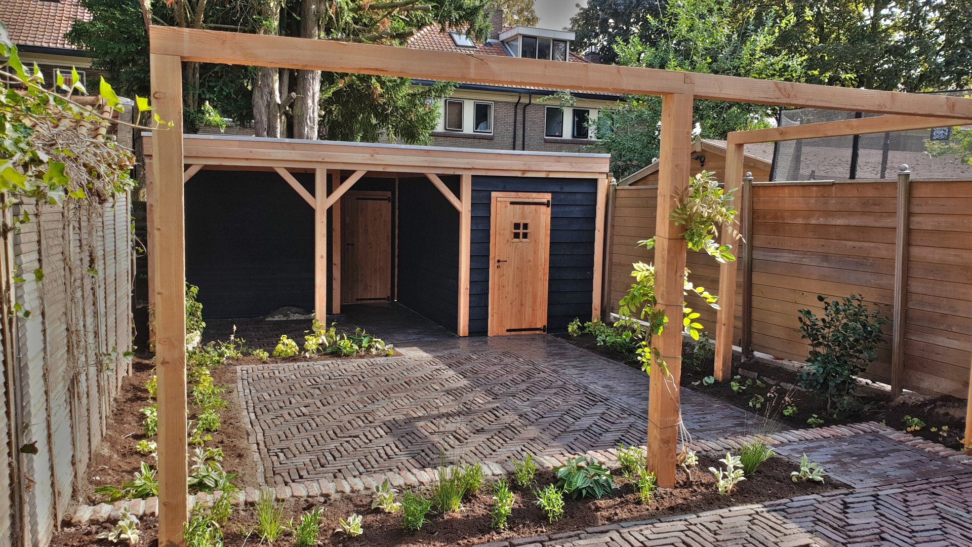 Houten schuur met een lichte houten deur, onder een houten pergola, omgeven door een betegeld terras en een schutting.