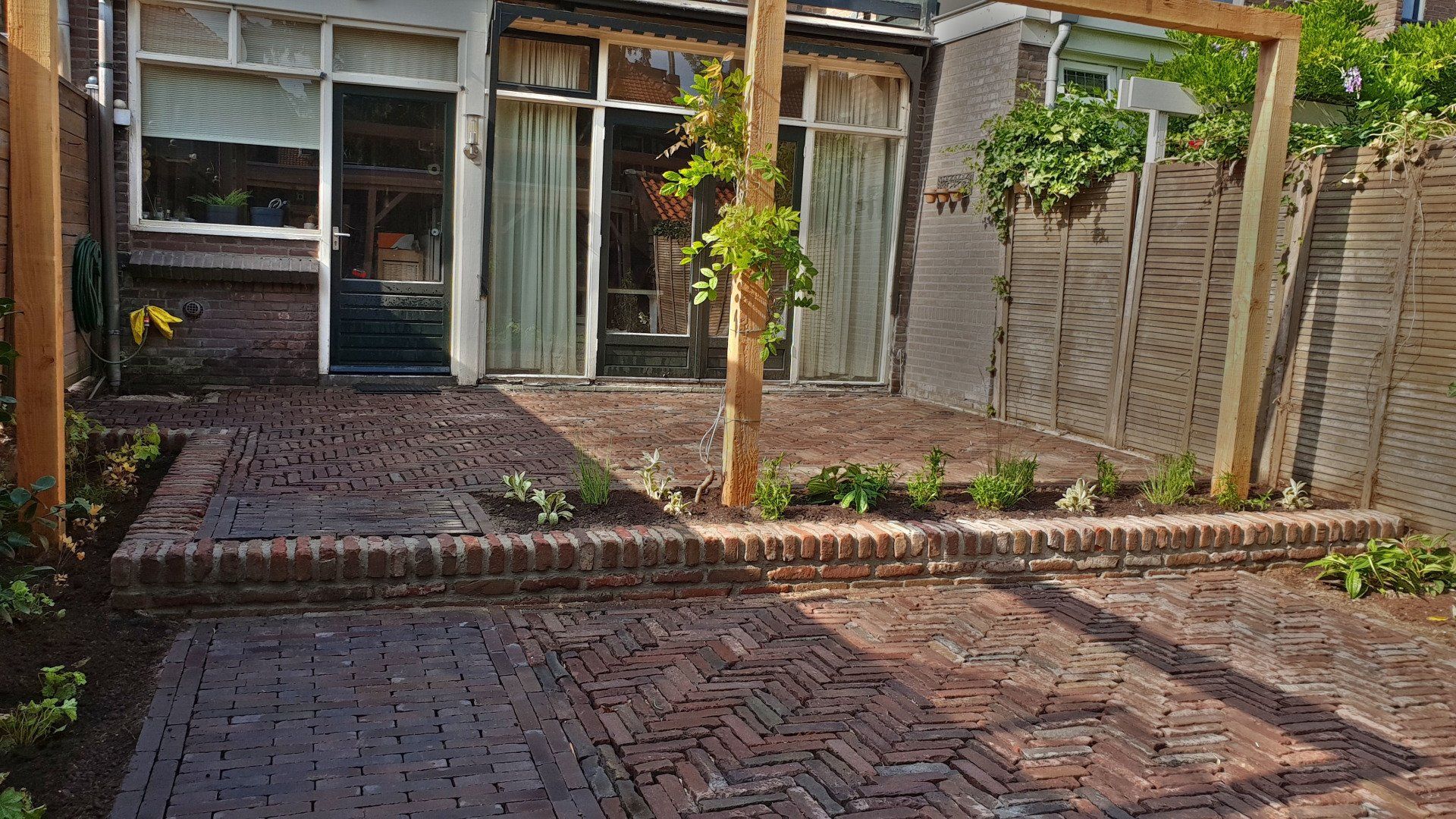 Bakstenen terras met houten pergola, plantenbed en huis met glazen deuren.