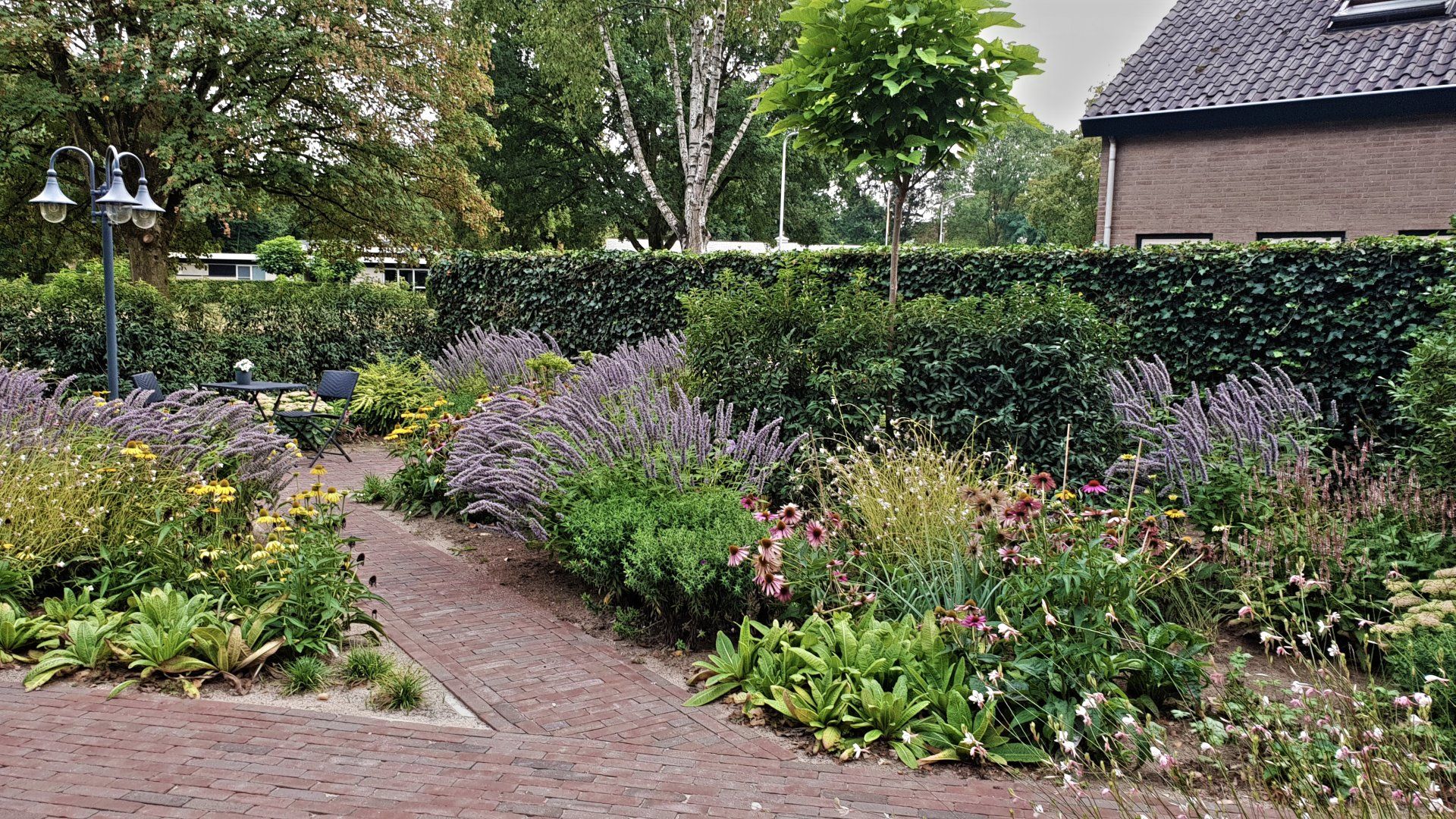 Een bakstenen pad loopt door een tuin vol paarse en gele bloemen en een hoge groene haag.