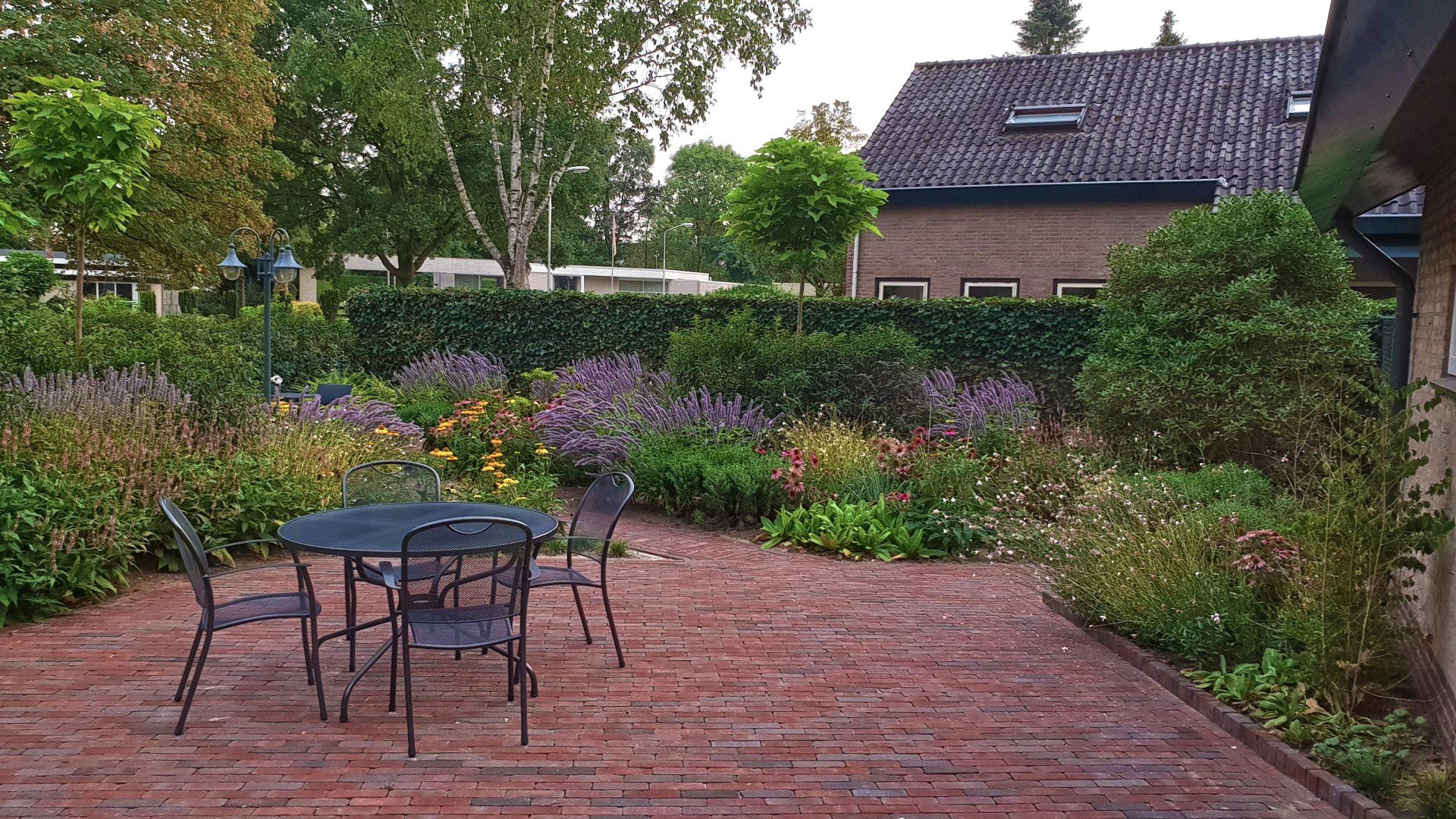 Een stenen terras met een zwarte tafel en stoelen, omgeven door een kleurrijke tuin en groene hagen, naast een stenen huis.