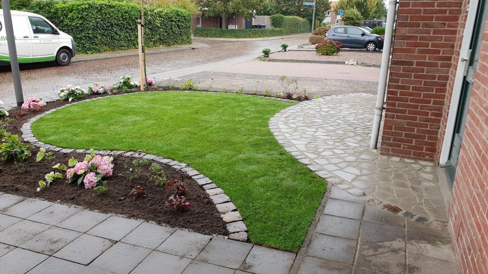 Kleine voortuin met groen gras, bloemperk en bakstenen rand. Kasseienpad en bakstenen gebouw zichtbaar.