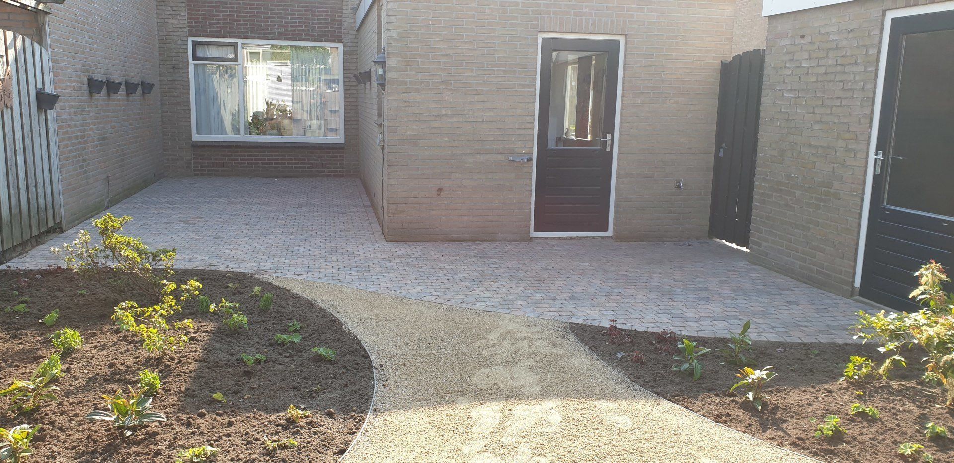 Een klein bakstenen gebouw met een geplaveide oprit en bloembedden met jonge planten.