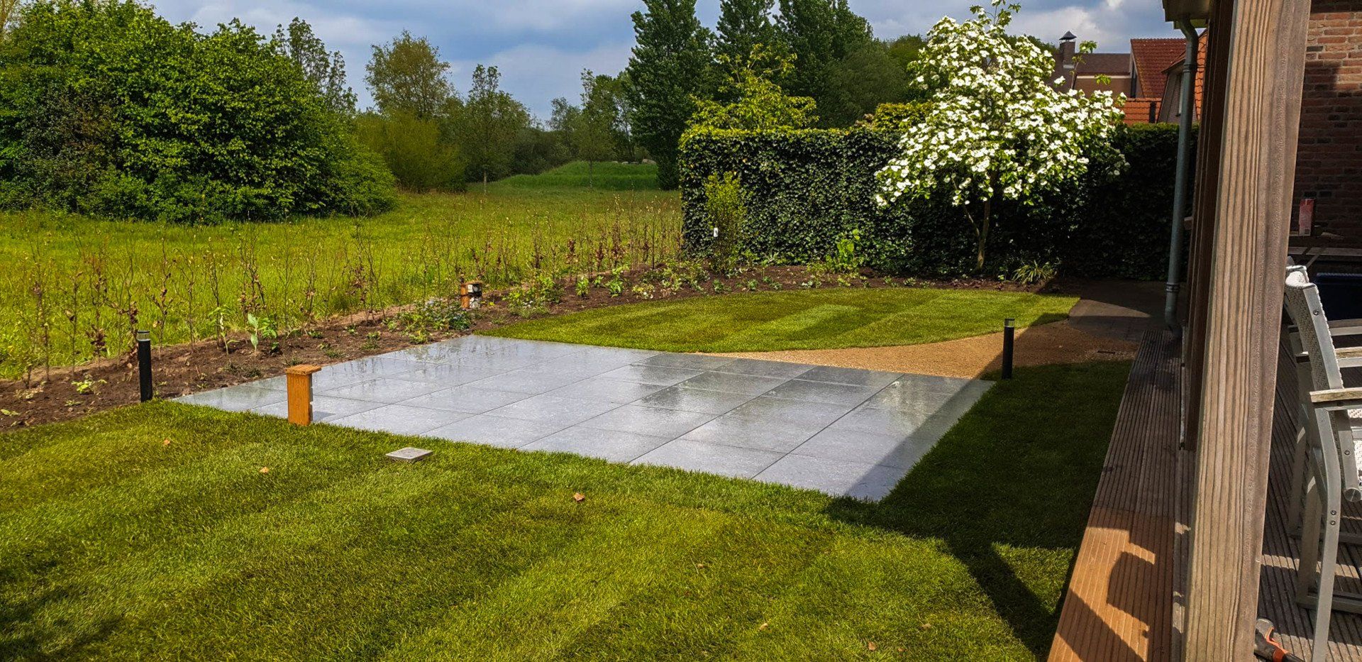 Een achtertuin met een rechthoekige betonnen plaat, gras en omringende begroeiing.
