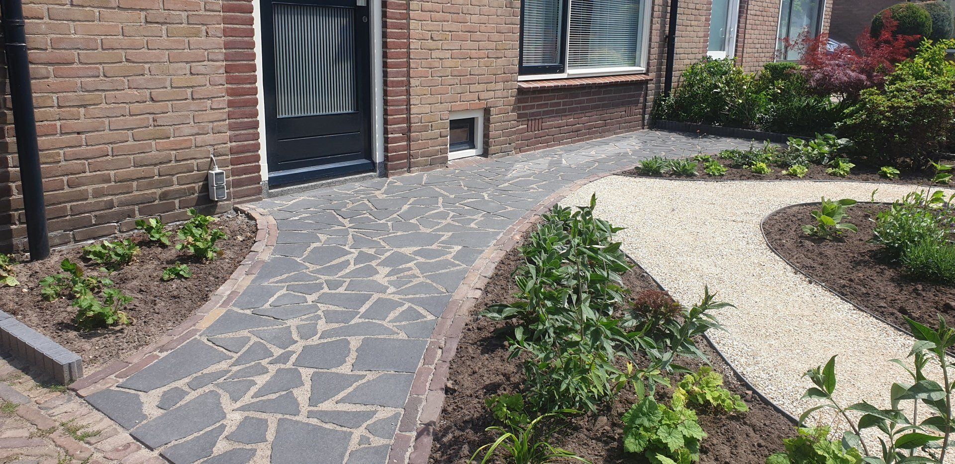 Een stenen pad leidt naar een voordeur en slingert langs bloemperken en een grindpad. Op de achtergrond is een bakstenen gebouw te zien.