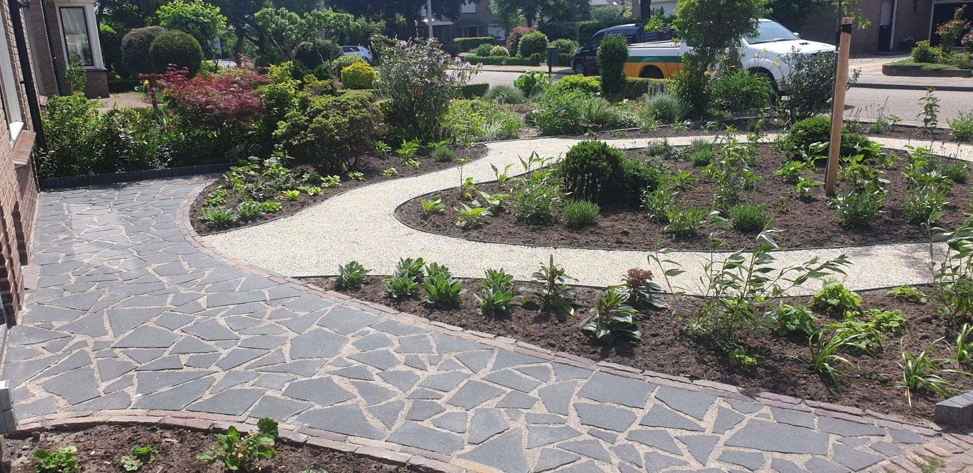 Een stenen pad slingert door een tuin met diverse planten, struiken en een centraal cirkelvormig bloembed.