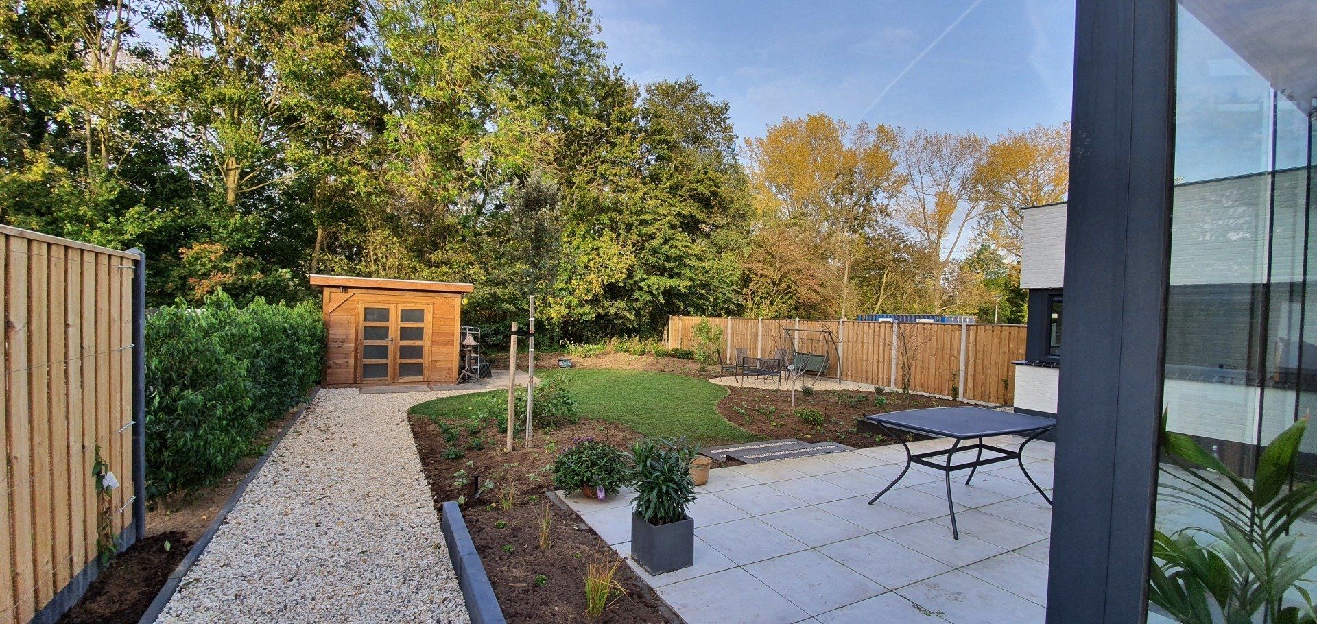 Een achtertuin met een stenen pad, schuur, terras, tafel en houten schutting, omgeven door bomen en groen.