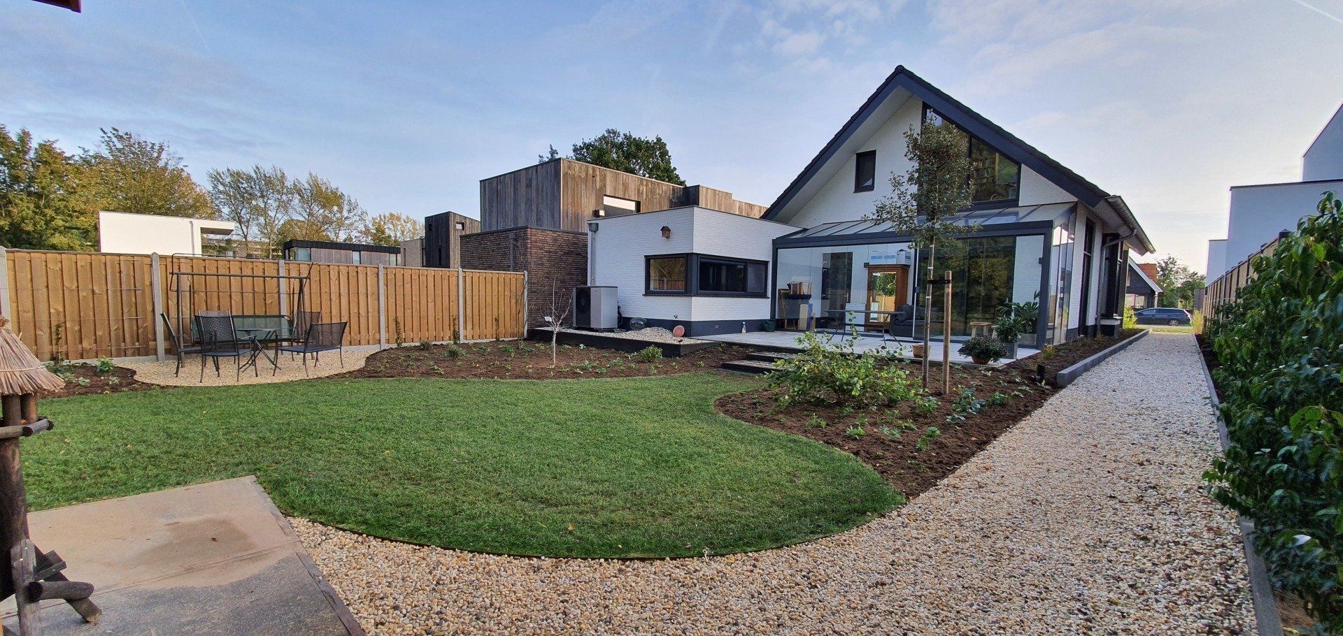 Een huis met een tuin voorzien van een grindpad, groen gras en een houten schutting. Een zonnige dag.