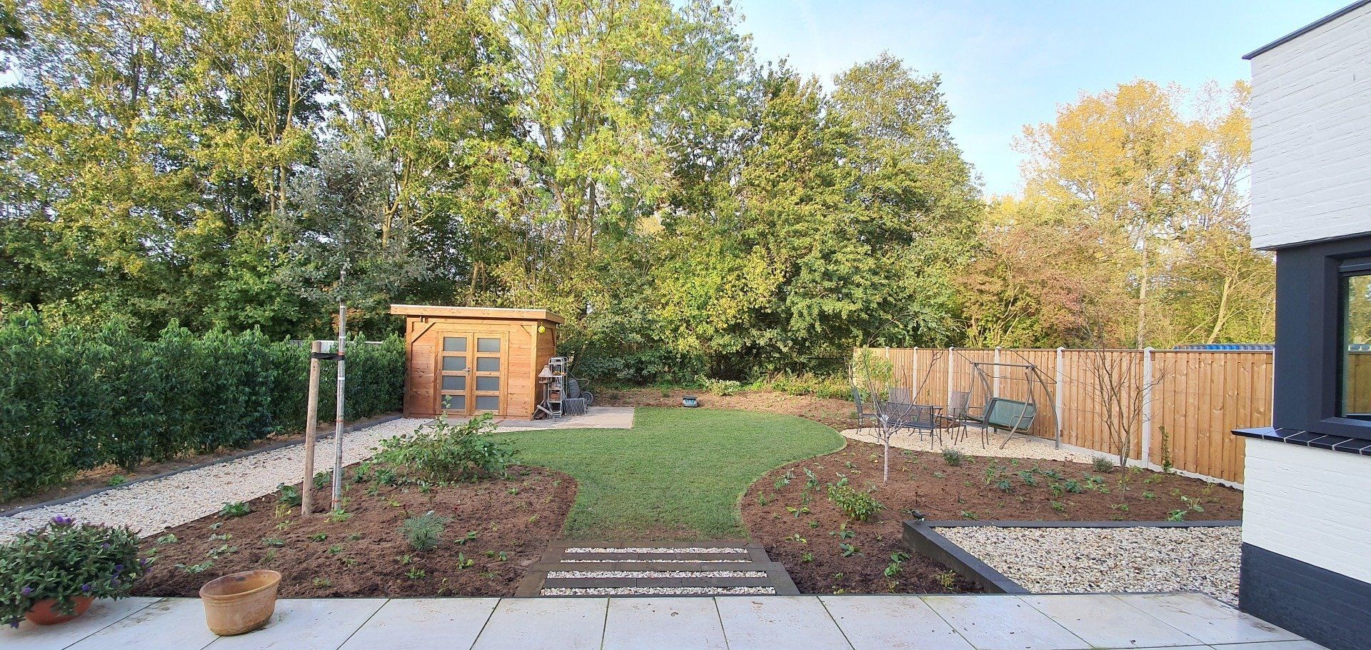 Een achtertuin met een houten schuur, een bestraat pad en groen gras, omgeven door bomen en een houten schutting.
