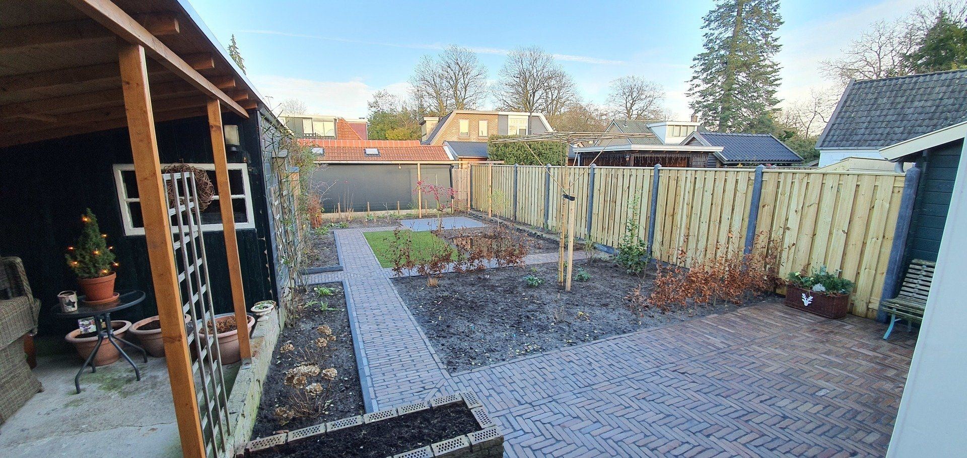 Een kleine achtertuin met een betegeld terras en een pad dat naar een tuin leidt. Er zijn houten schuttingen en een schuur aanwezig.