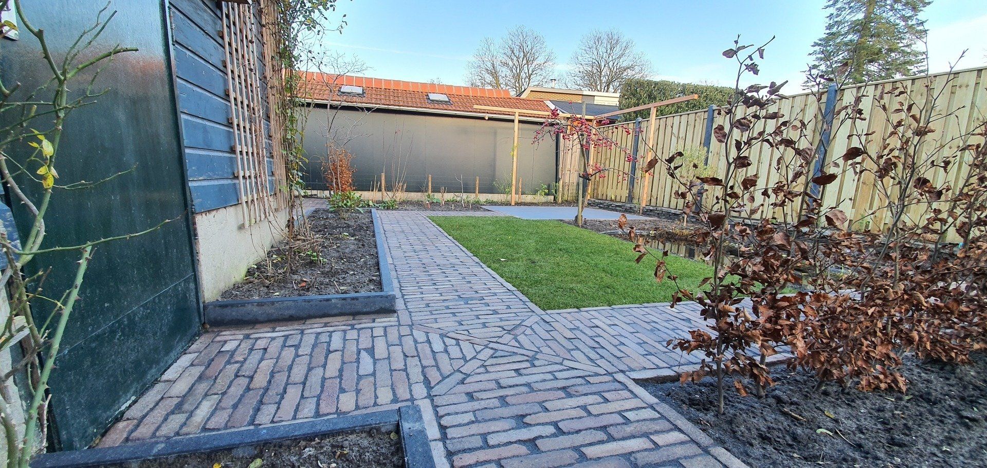 Een bakstenen pad leidt door een tuin met een groen gazon en een houten schutting onder een heldere hemel.