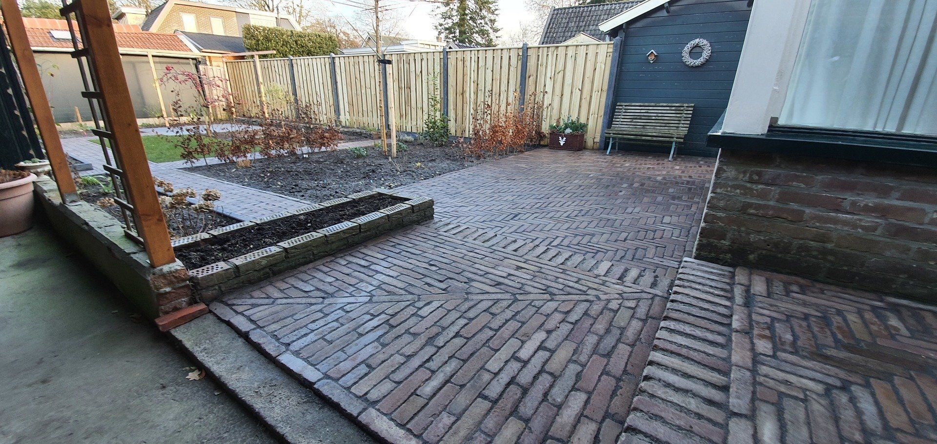 Een stenen terras met verhoogde plantenbak, een houten schutting en een schuur in de achtertuin.