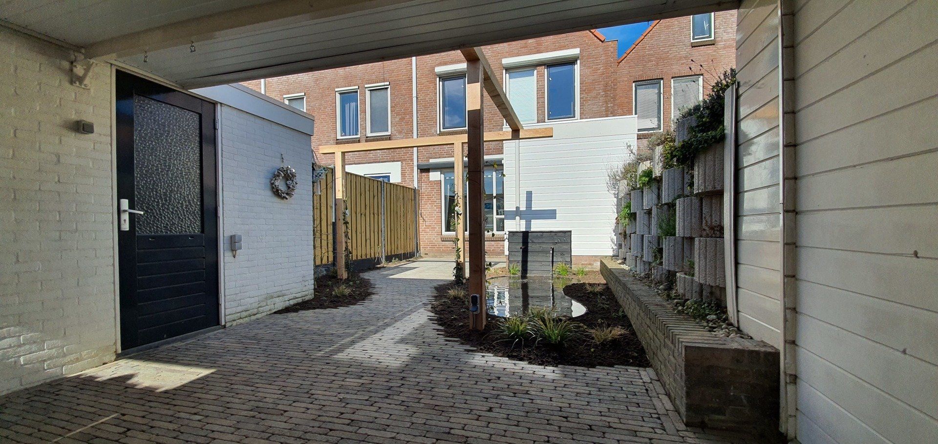 Pad dat naar een achtertuin leidt. Zwarte deur links, tuinmuur met planten rechts. Houten schutting en gebouwen op de achtergrond.