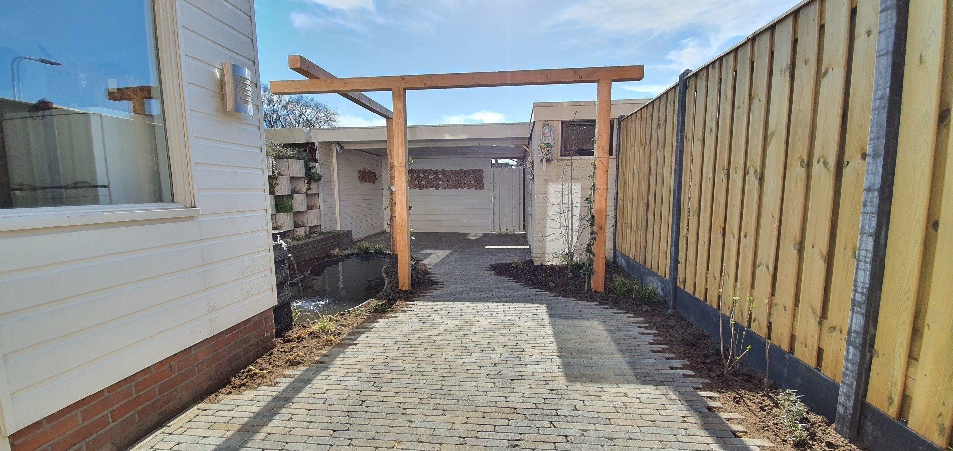Een geplaveid pad leidt naar een houten boog en een kleine binnenplaats. Rechts staat een houten hek, links een wit gebouw.