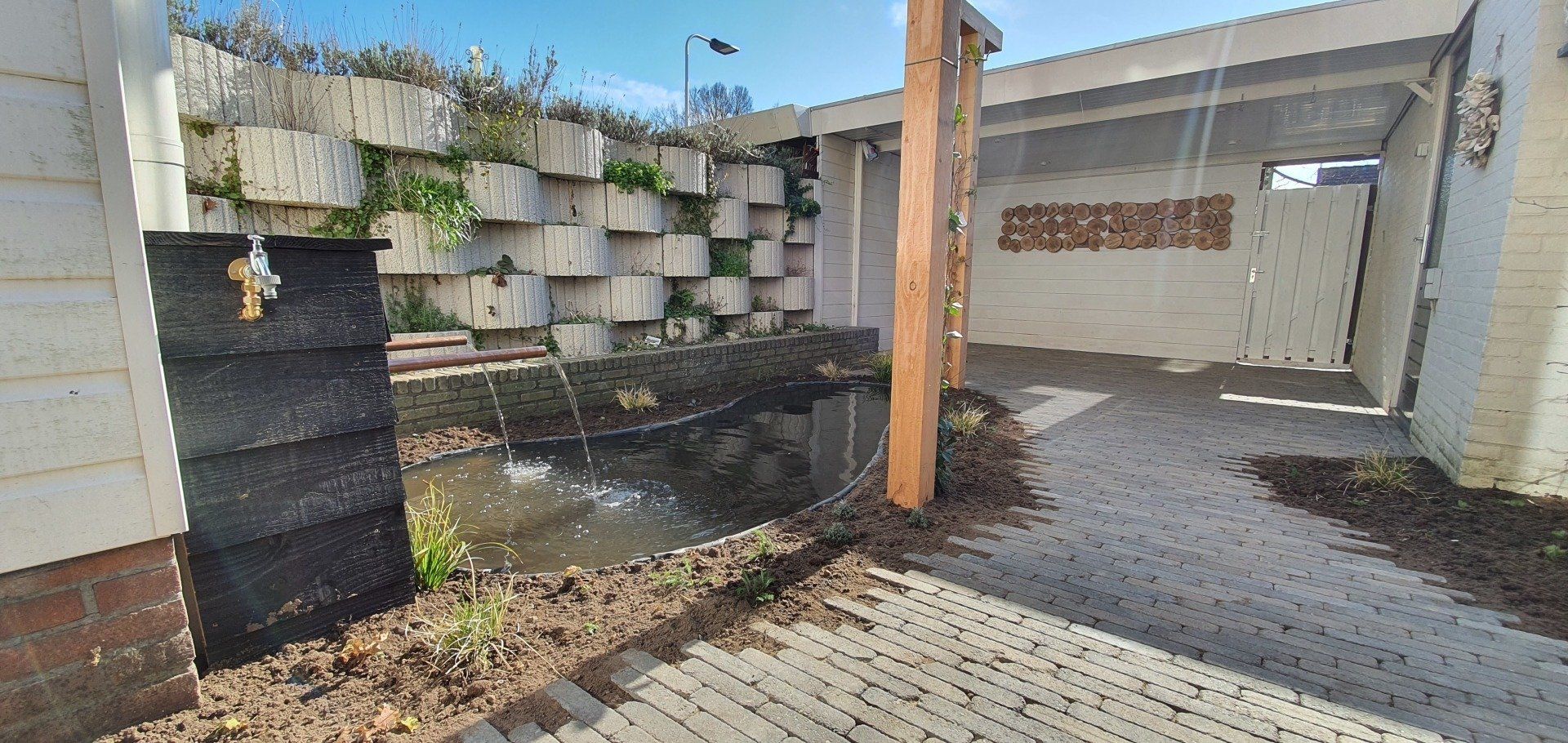 Een tafereel aan het water met een gestapelde keermuur en een carport. Een zwarte watercontainer heeft een kraan.
