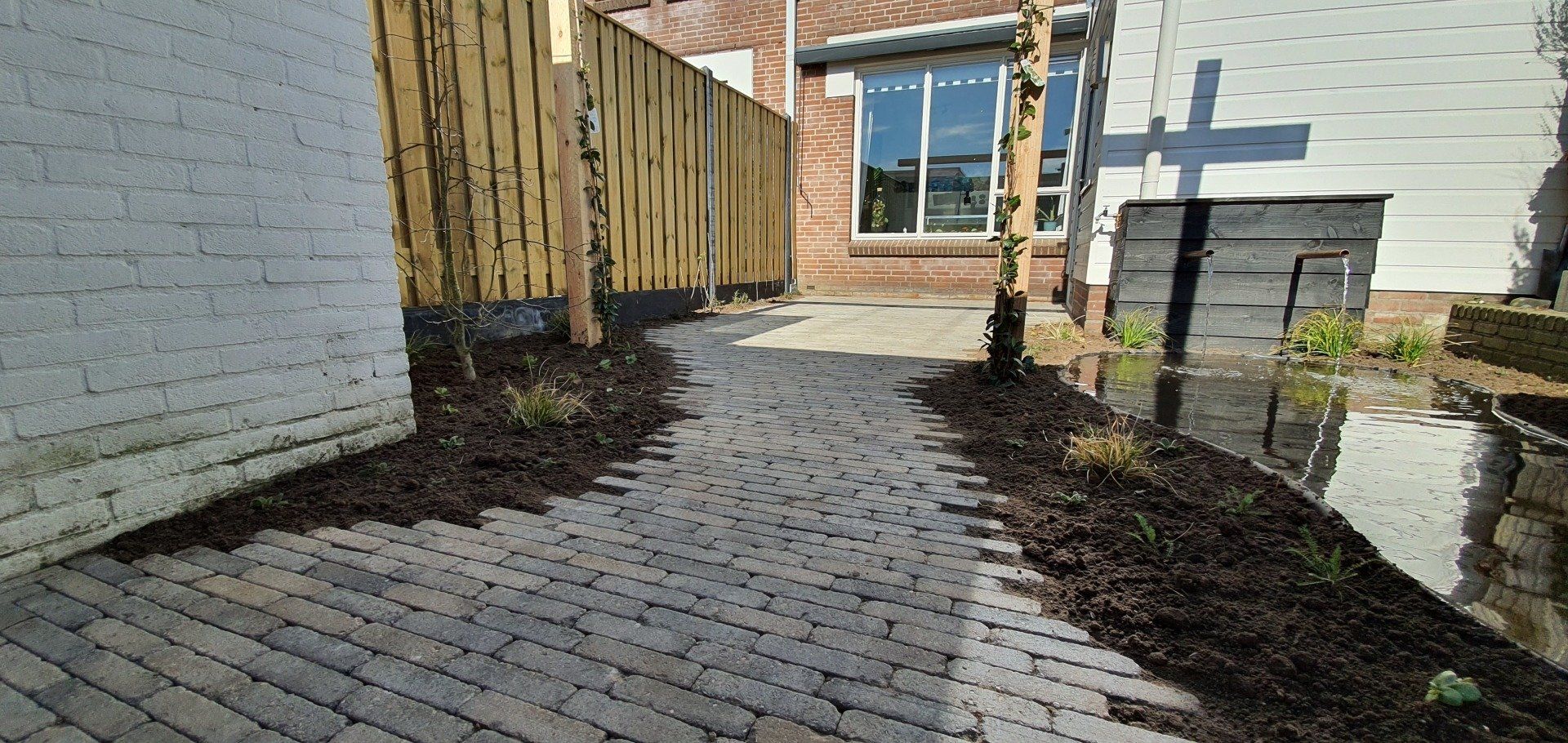 Een bakstenen pad kronkelt door een aangelegde tuin, langs een houten schutting en een bijgebouw.