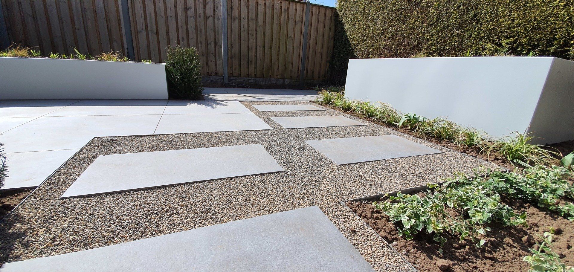 Een moderne tuin met stenen bestrating, grind en rechthoekige plantenbakken gevuld met groen. Houten schutting op de achtergrond.