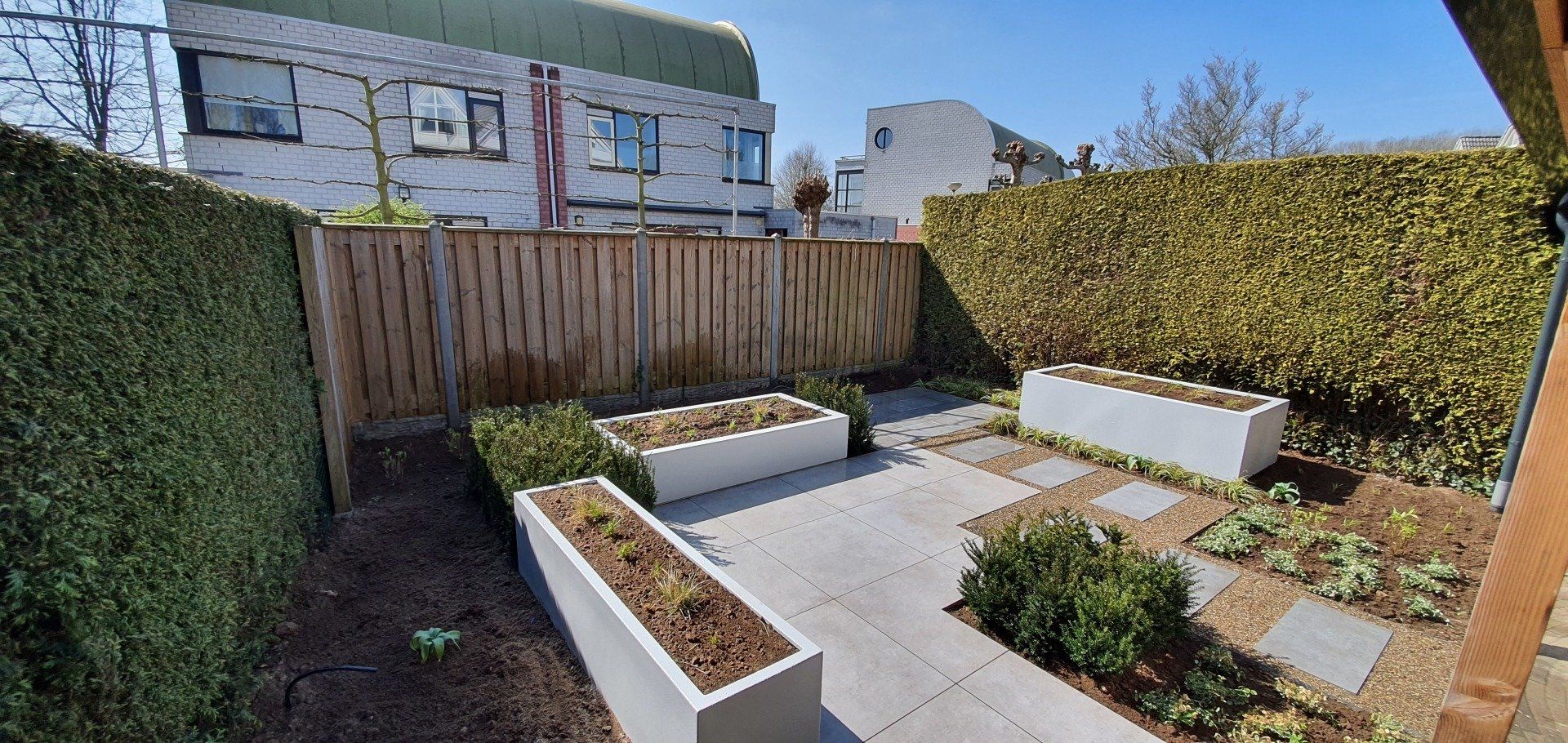 Een kleine, moderne achtertuin met betonnen plantenbakken en natuurstenen tegels, omgeven door een houten schutting en hagen.