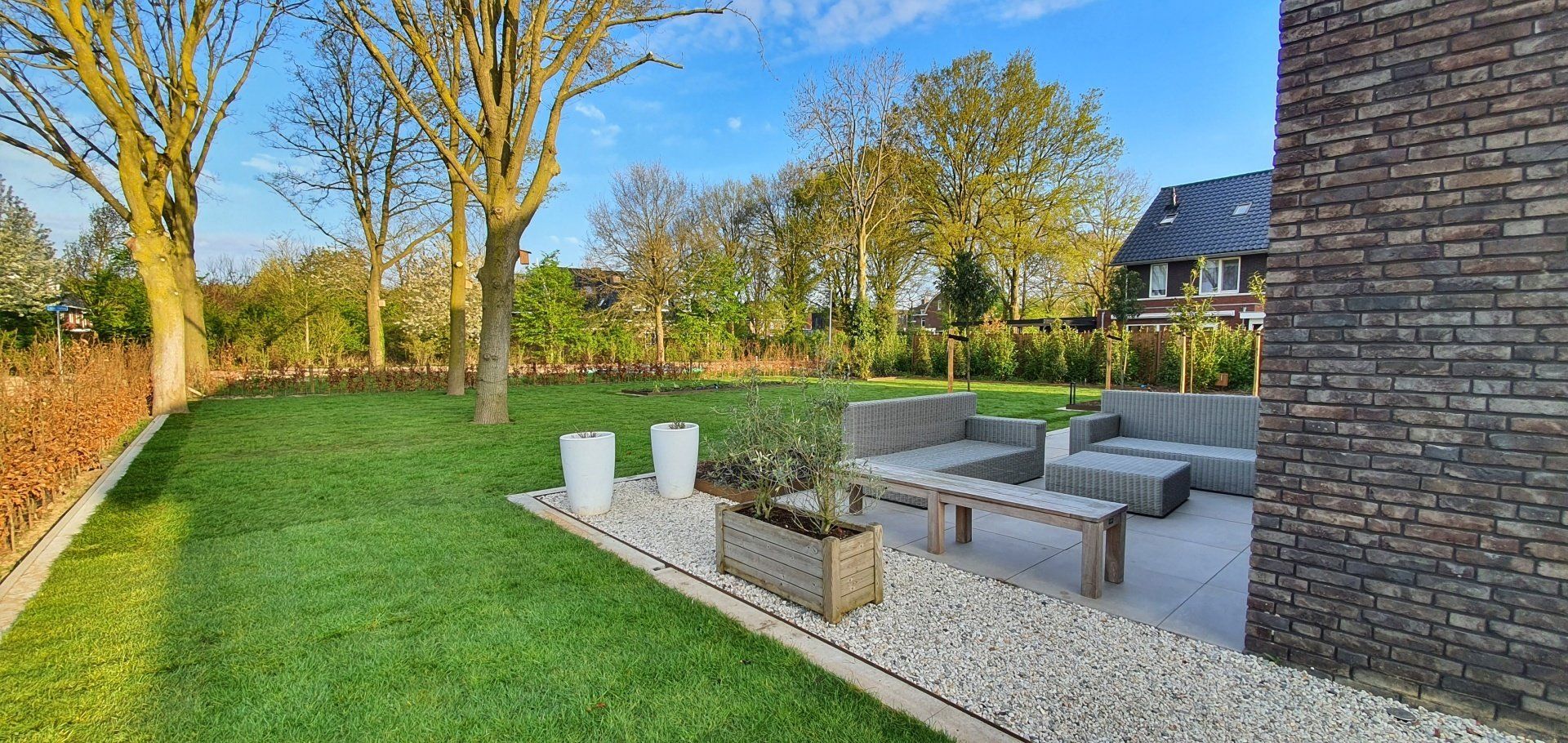 Een achtertuin met gazon, bomen en tuinmeubilair naast een bakstenen muur onder een blauwe hemel.