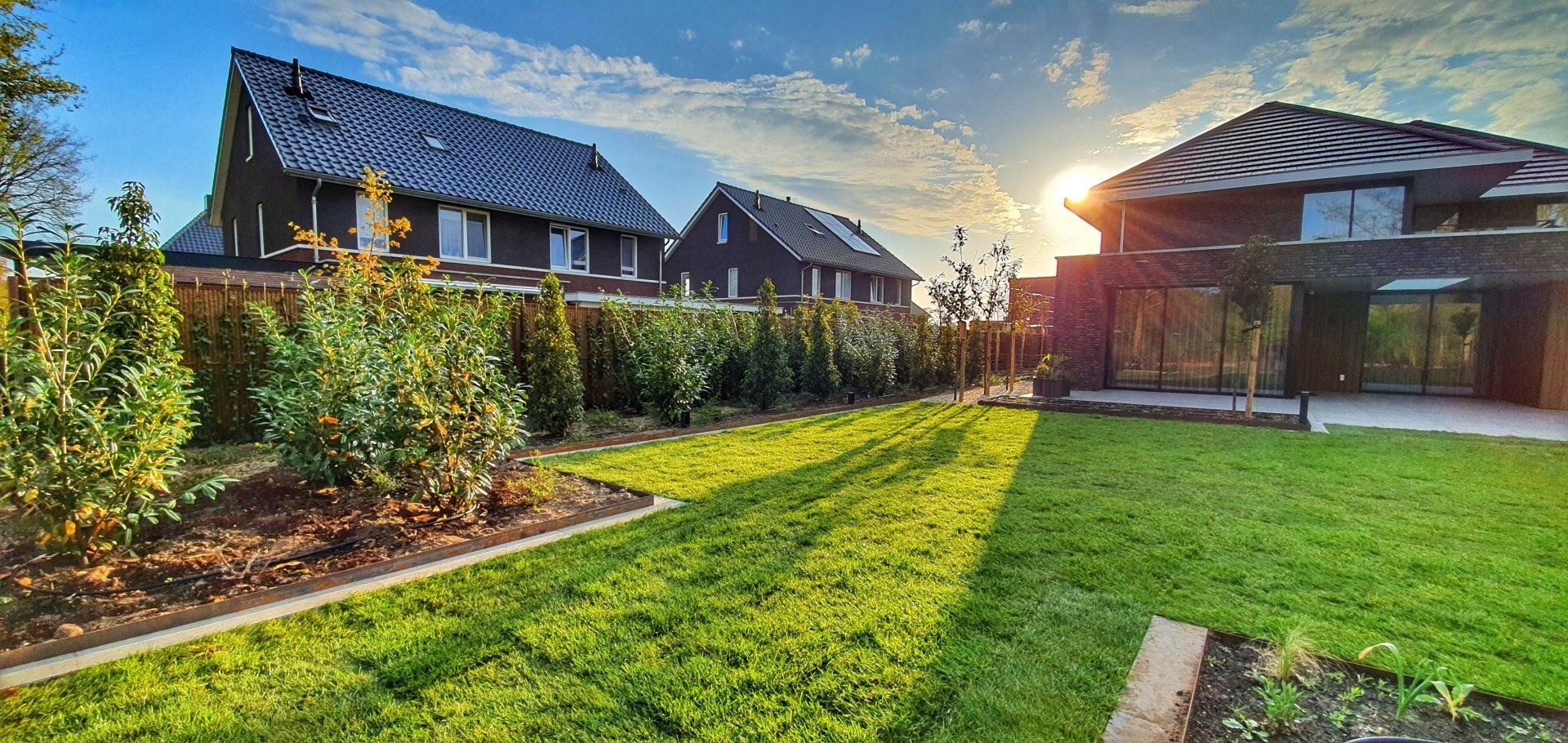 Een weelderig groen gazon met keurig gesnoeide struiken en moderne huizen. De zon schijnt fel.