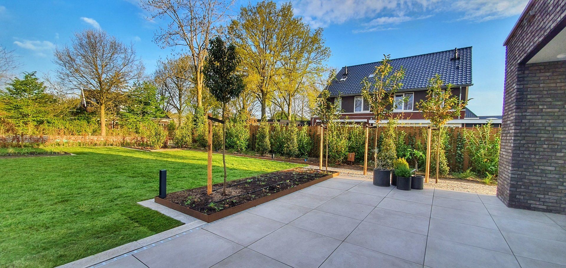 Terras met groen gazon en bomen, een huis op de achtergrond. Blauwe lucht erboven.