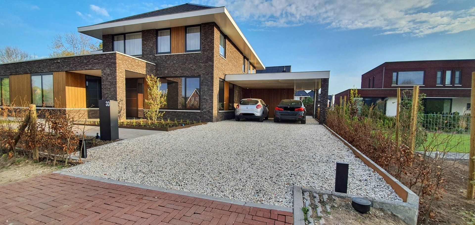 Een modern, twee verdiepingen tellend bakstenen huis met een oprit van grind en twee geparkeerde auto's onder een carport.