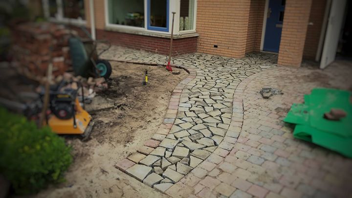 Aanleg van een pad in volle gang; kasseien worden gelegd langs een gebogen pad voor een huis, omringd door gereedschap en materialen.