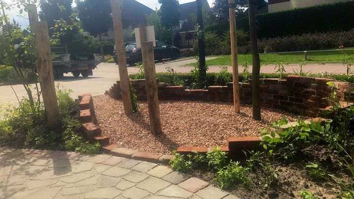 Een kleine ronde tuin met bakstenen randen en houten palen, gevuld met houtsnippers; een geplaveid pad leidt ernaartoe.