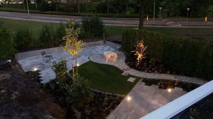 Een terras in de achtertuin met buitenverlichting, bomen, gazon en een stenen pad.