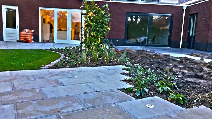 Terras in de achtertuin met stenen bestrating, bloemperken, gazon en een huis met glazen deuren.