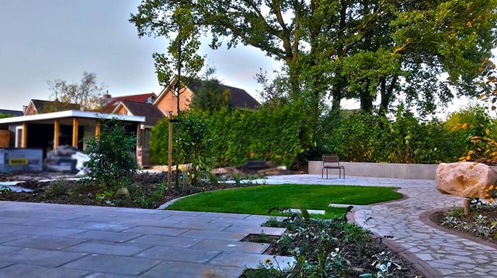 Stenen terras en pad in een aangelegde tuin met gazon, struiken en bomen; een bankje in de verte.
