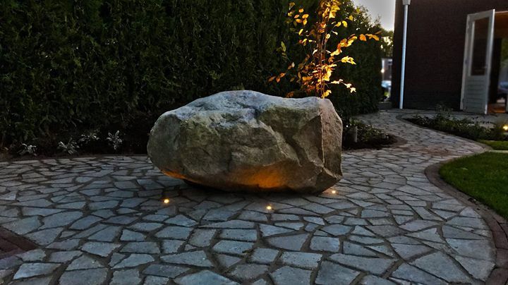Een grote rotssculptuur, verlicht door grondspots, staat op een stenen pad, met groene struiken op de achtergrond.