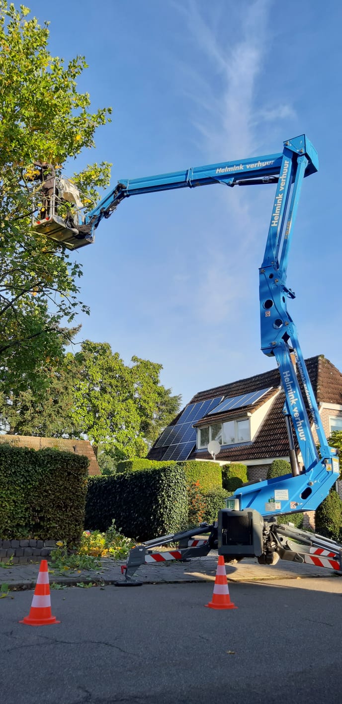 Een blauwe hoogwerker snoeit een boom. Oranje kegels blokkeren de weg. Op de achtergrond is een huis te zien.