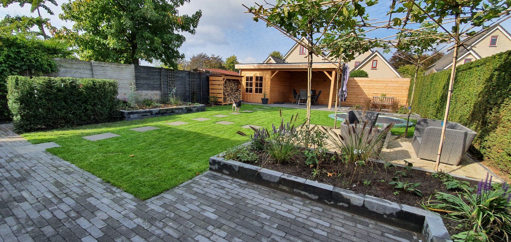 Een achtertuin met een houten constructie, een stenen terras, gras en bomen. Een zonnige dag.