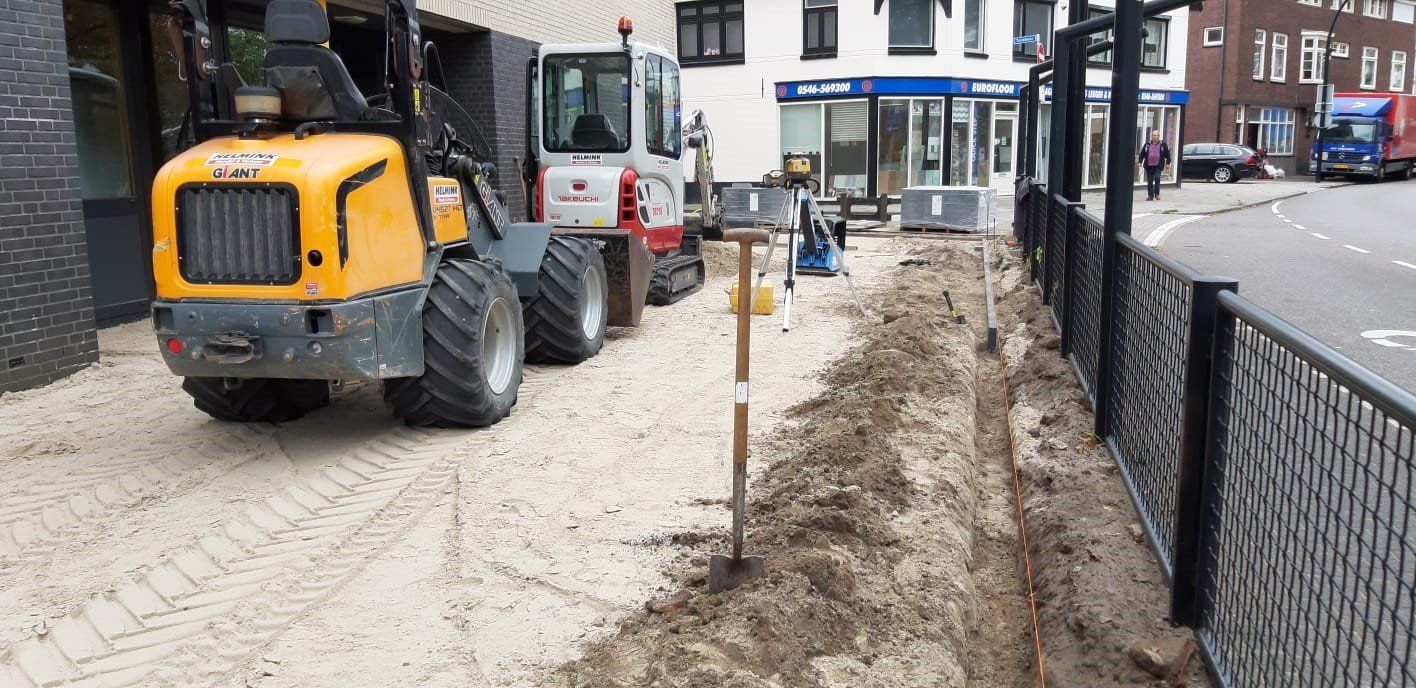 Bouwterrein met zware machines, een greppel en een hek naast een straat met gebouwen.