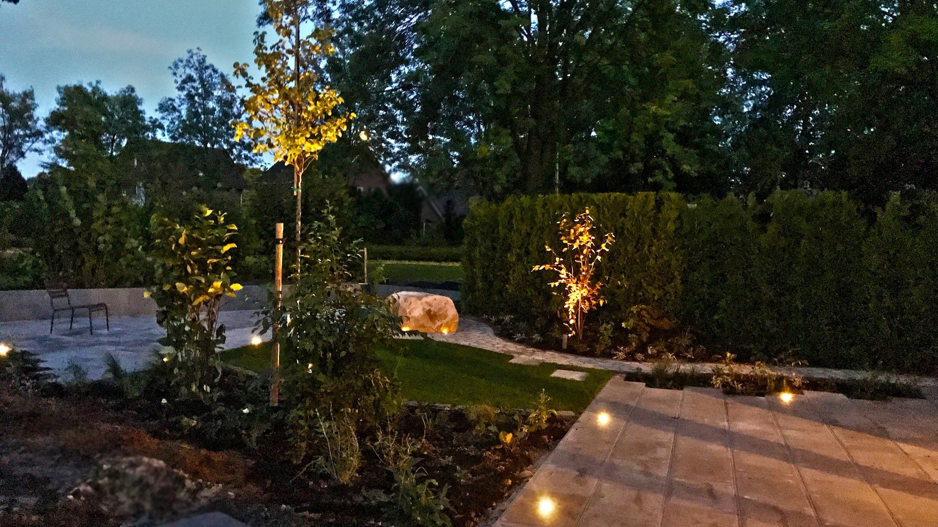 Tuin bij schemering met bomen, hagen, verlichte paden en decoratieve verlichting.