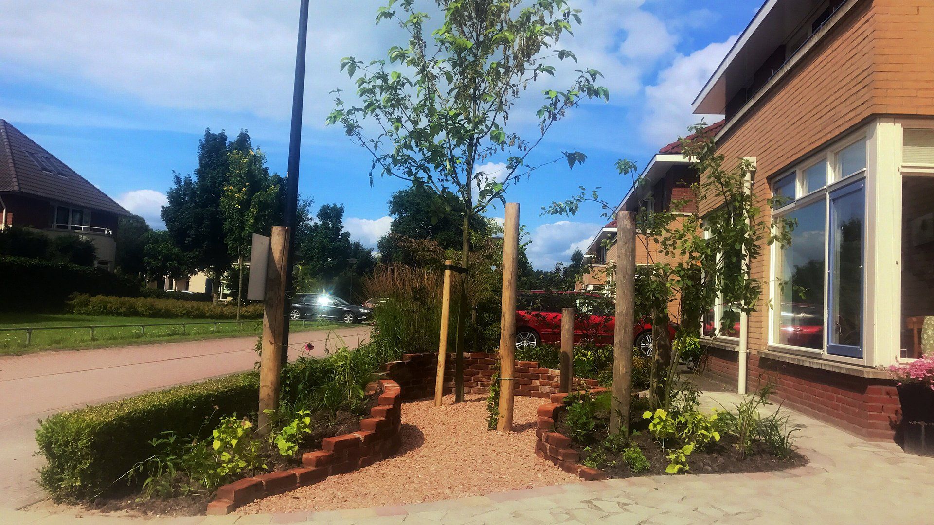 Een tuintje langs de straat met kleine boompjes, bruine bakstenen en een rode auto, vlakbij een bakstenen gebouw.